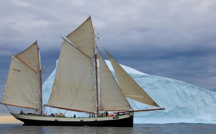 Traditional sailing ship Tecla will all her sails up, passing a large white iceberg in front of a moody sky. Polar exploration sailing adventure holidays with Classic Sailing.